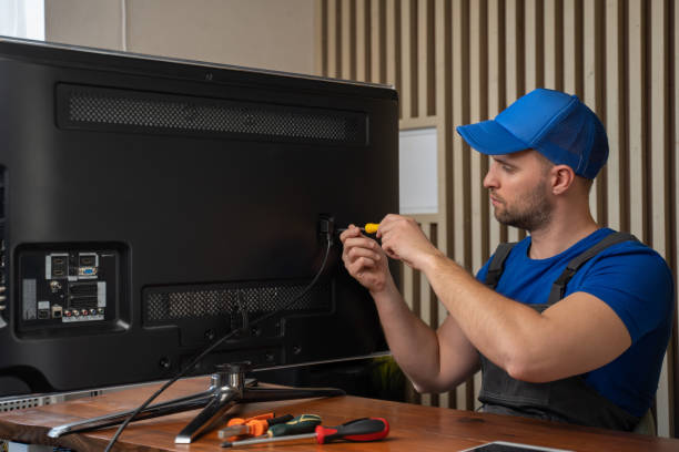 Master in blue T-shirt smiles after repairing TV set connector with screwdriver sitting at table. Diagnosis and repair of broken household appliances at home